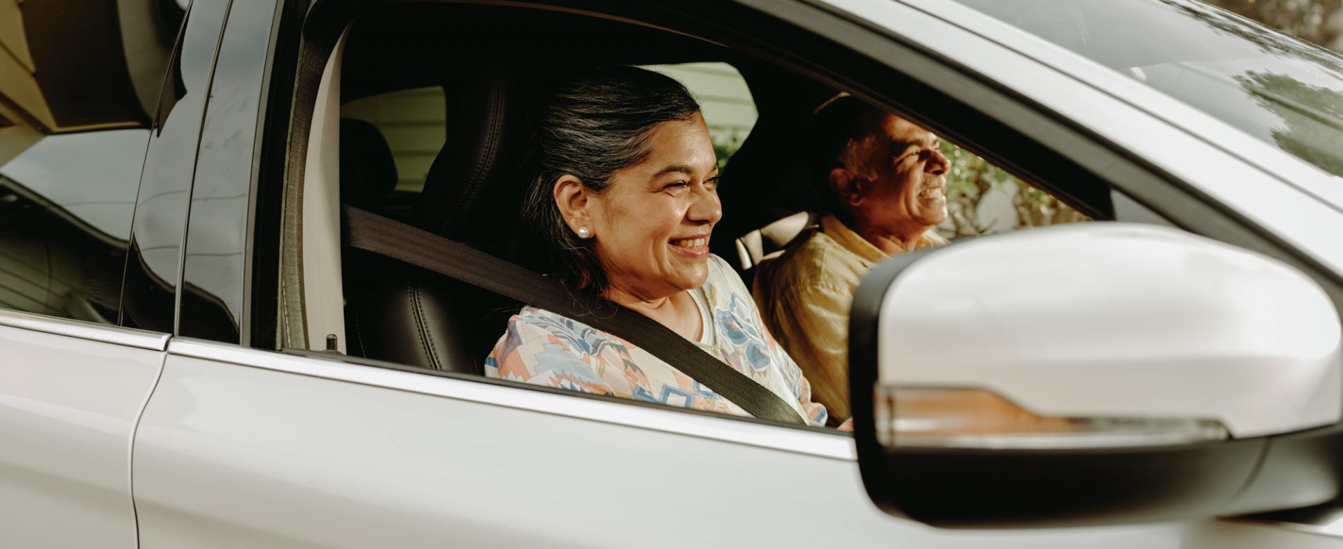 A woman is seen in the drivers seat of a white car she is holding the steering wheel and smiling. A male passenger sits in the front passenger seat