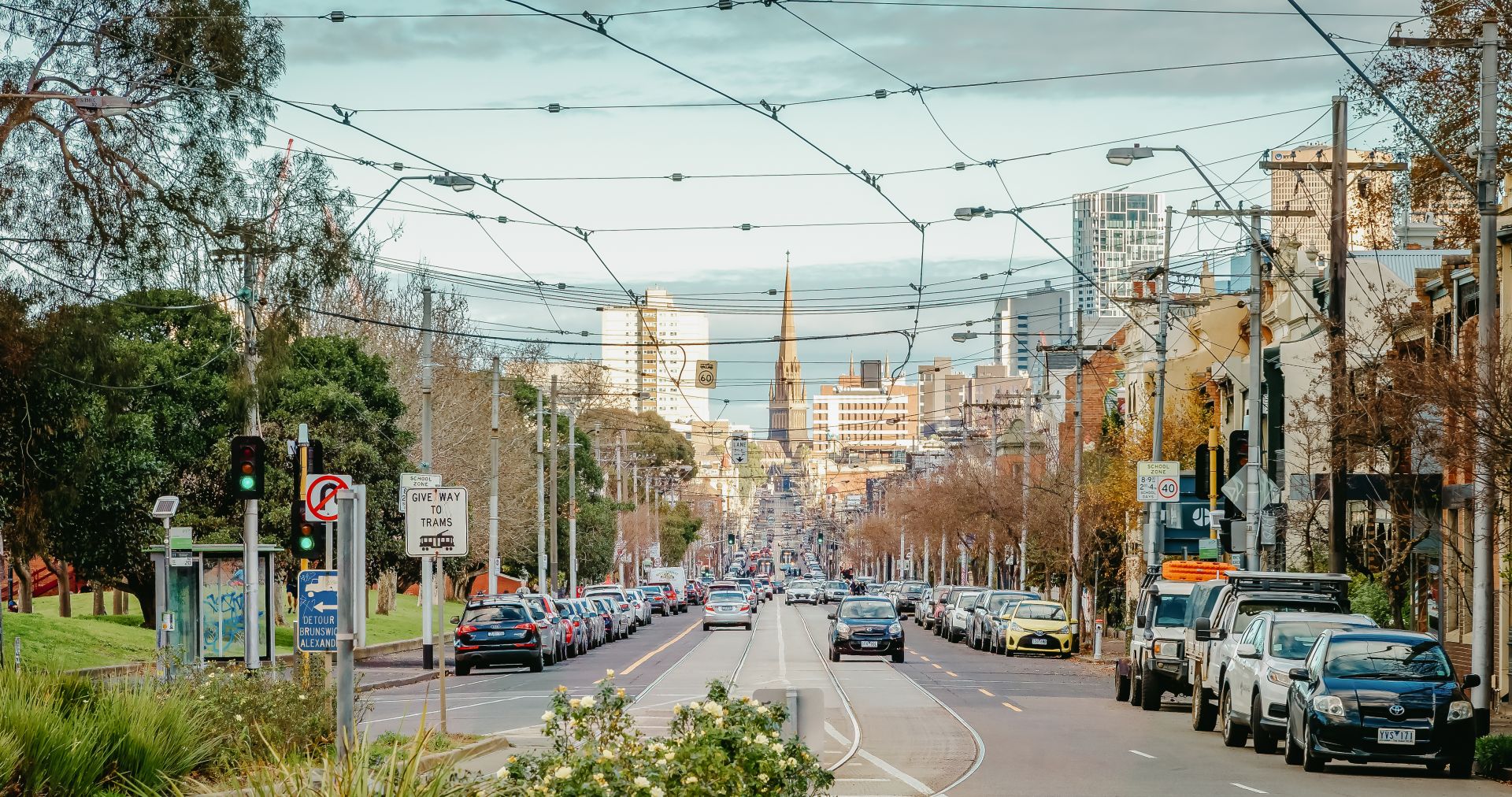 Image shows a road in Fitzroy Melbourne with tram tracklines and many cars seen parked on the road.