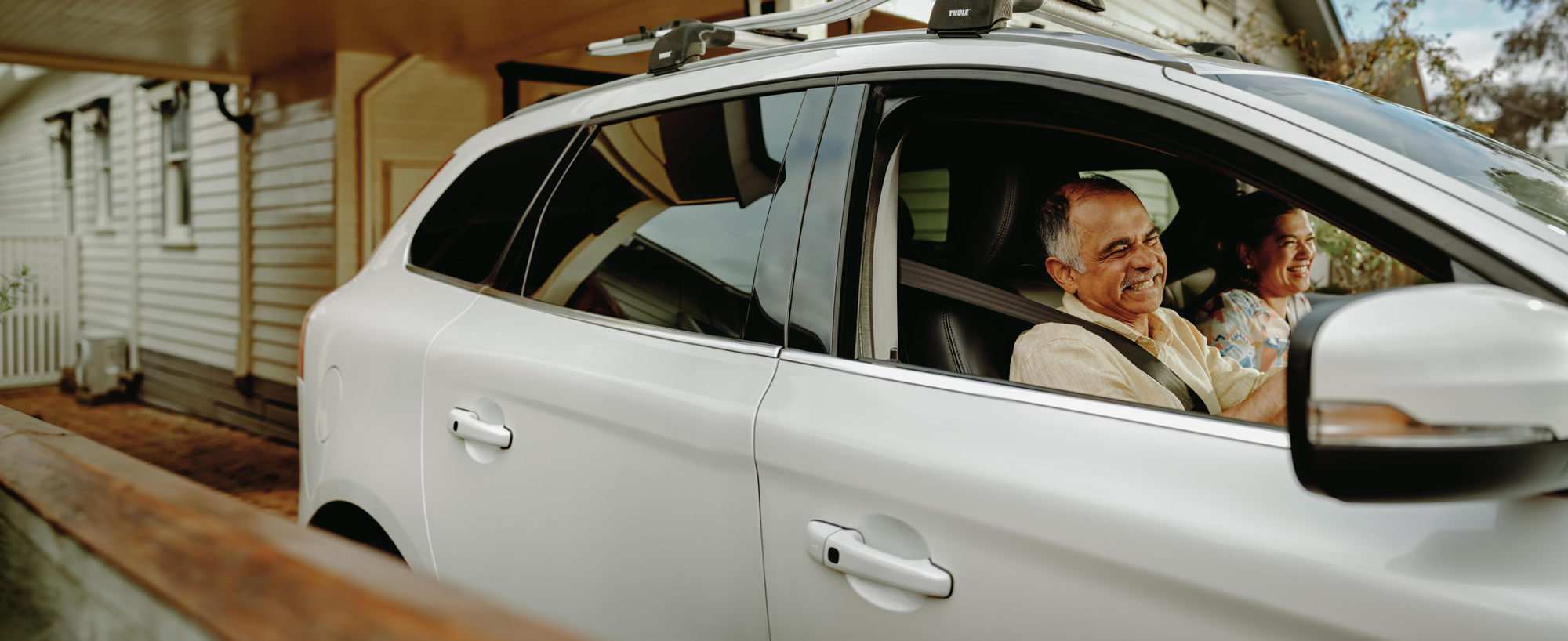 A man is seen in the drivers seat of a white car, we is holding the steering wheel smiling. A woman is seen in the front passenger seat