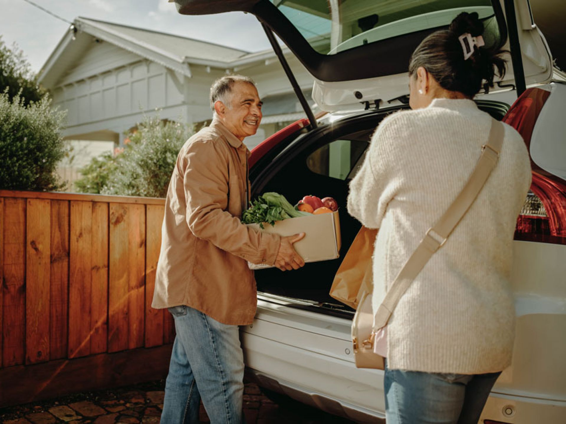 A man and a woman are seen removing a box of fresh produce from the boot of their white car. 