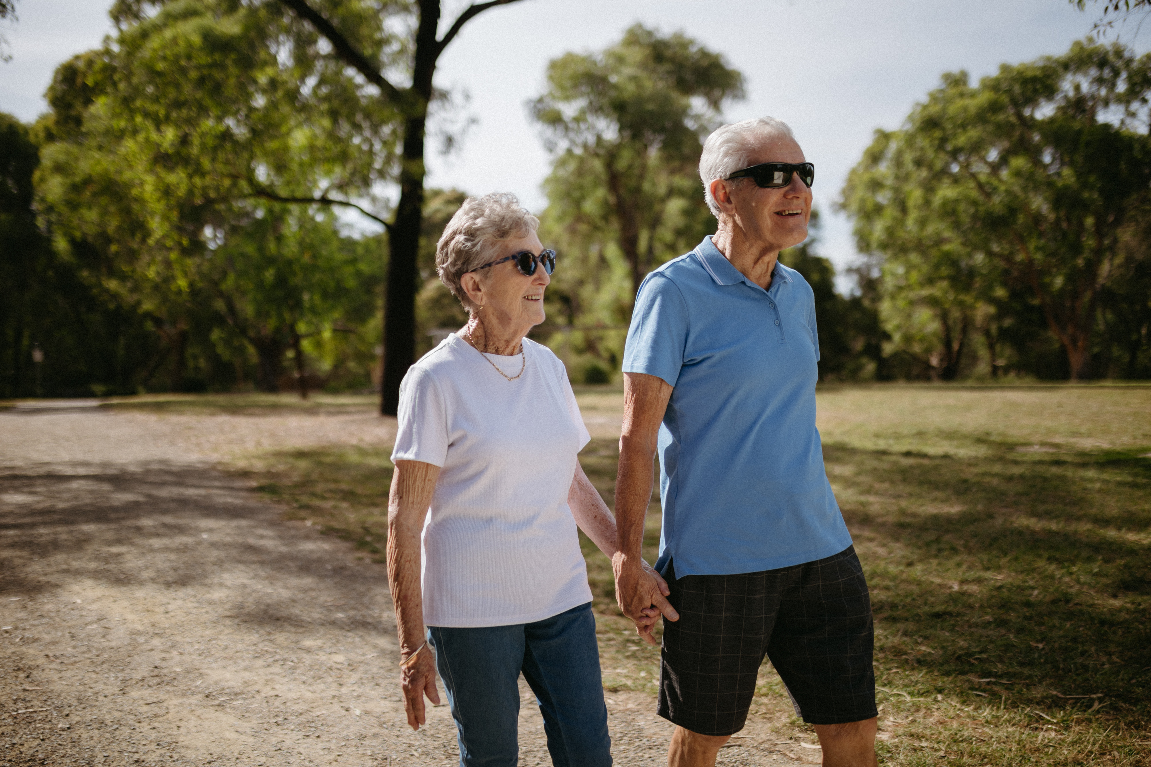 A man in his 80's wearing a blue top and a woman in her 80's wearing a white top are seen walking on a bush track