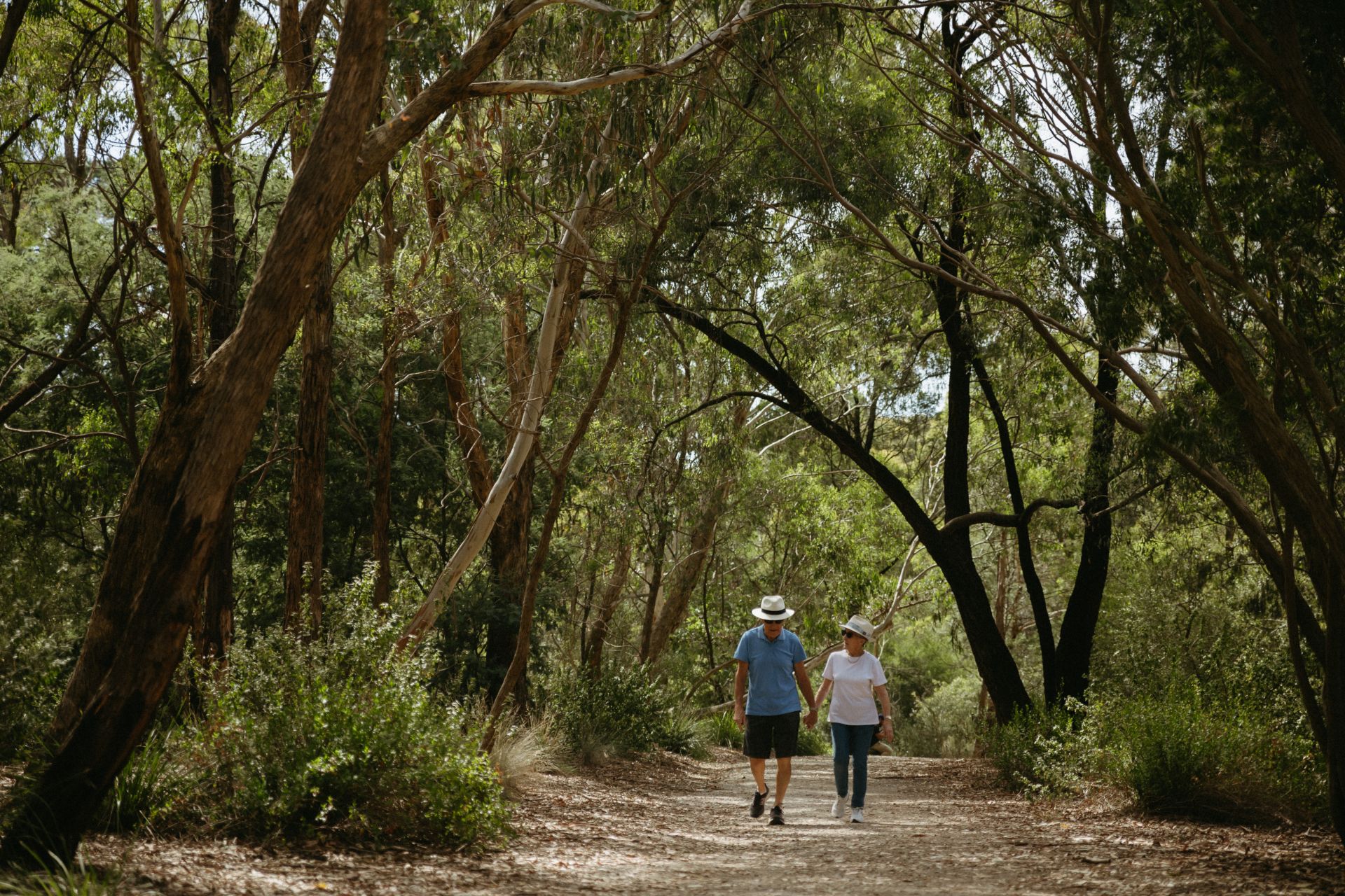 A man in a blue top and a light coloured hat is seen holding hands with a woman wearing a white top and light coloured hat as they walk on a bush track