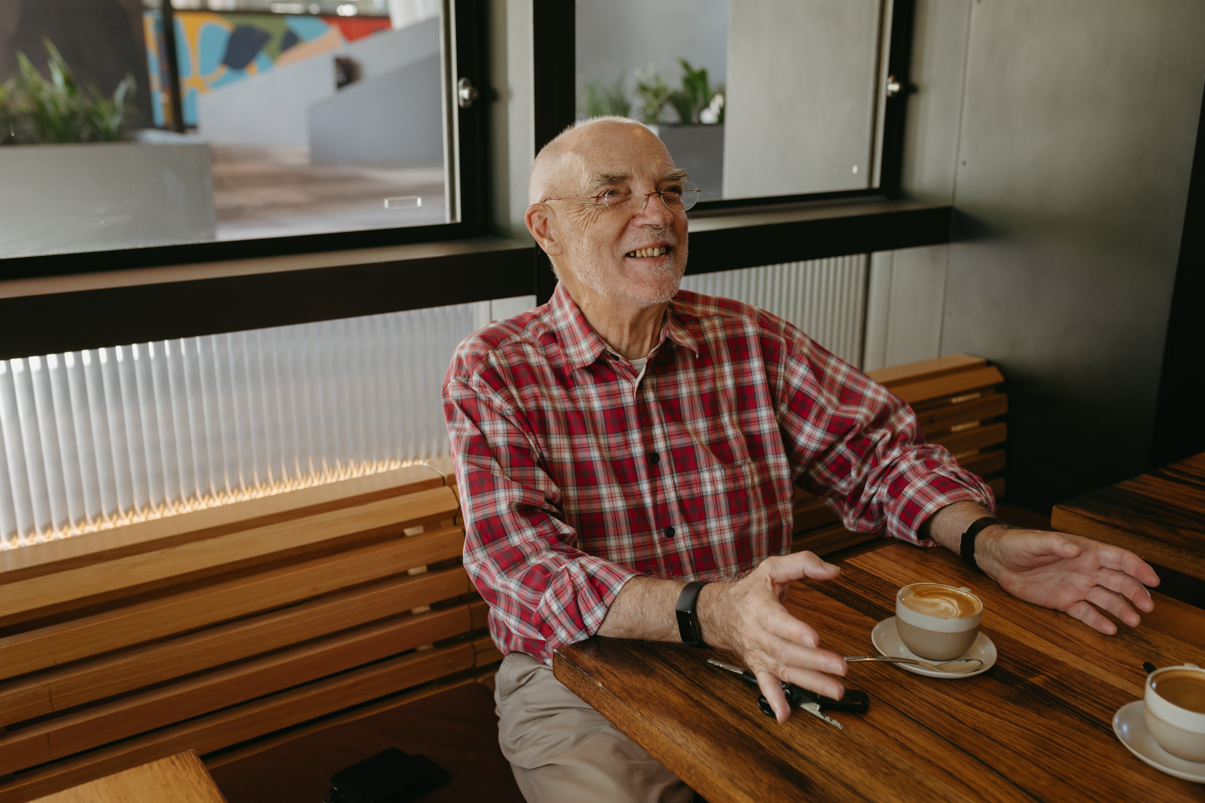 Older man in a red shirt seen sitting at a cafe with a coffee laughing