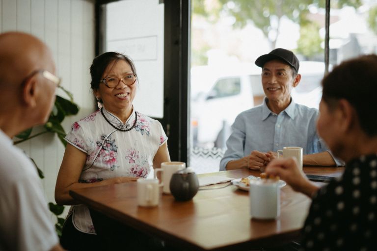 A man and woman are seen smiling whilst having a coffee with friends. The man is wearing a blue shirt and a black cap and the female is wearing a white top and glasses. 