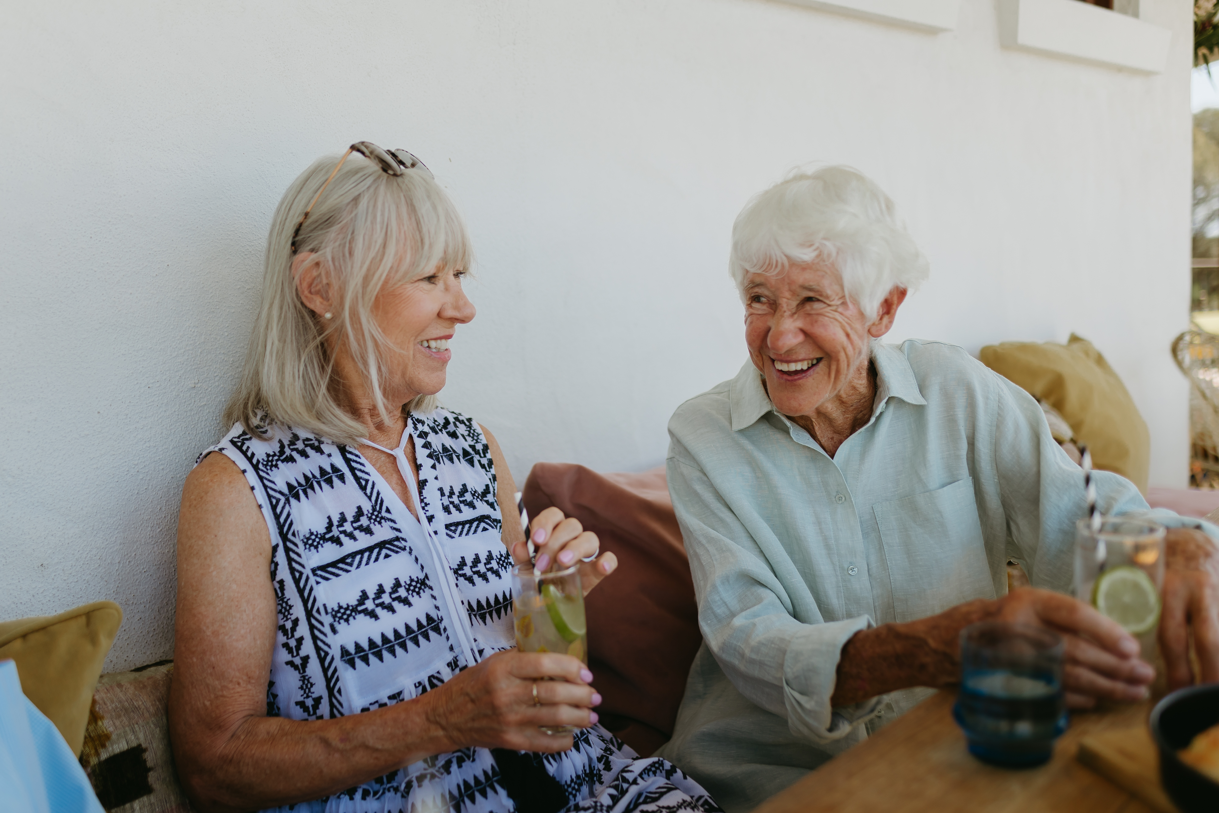 Two women in their 70's are seen smiling whilst sitting at a cafe table both holding drinks. 