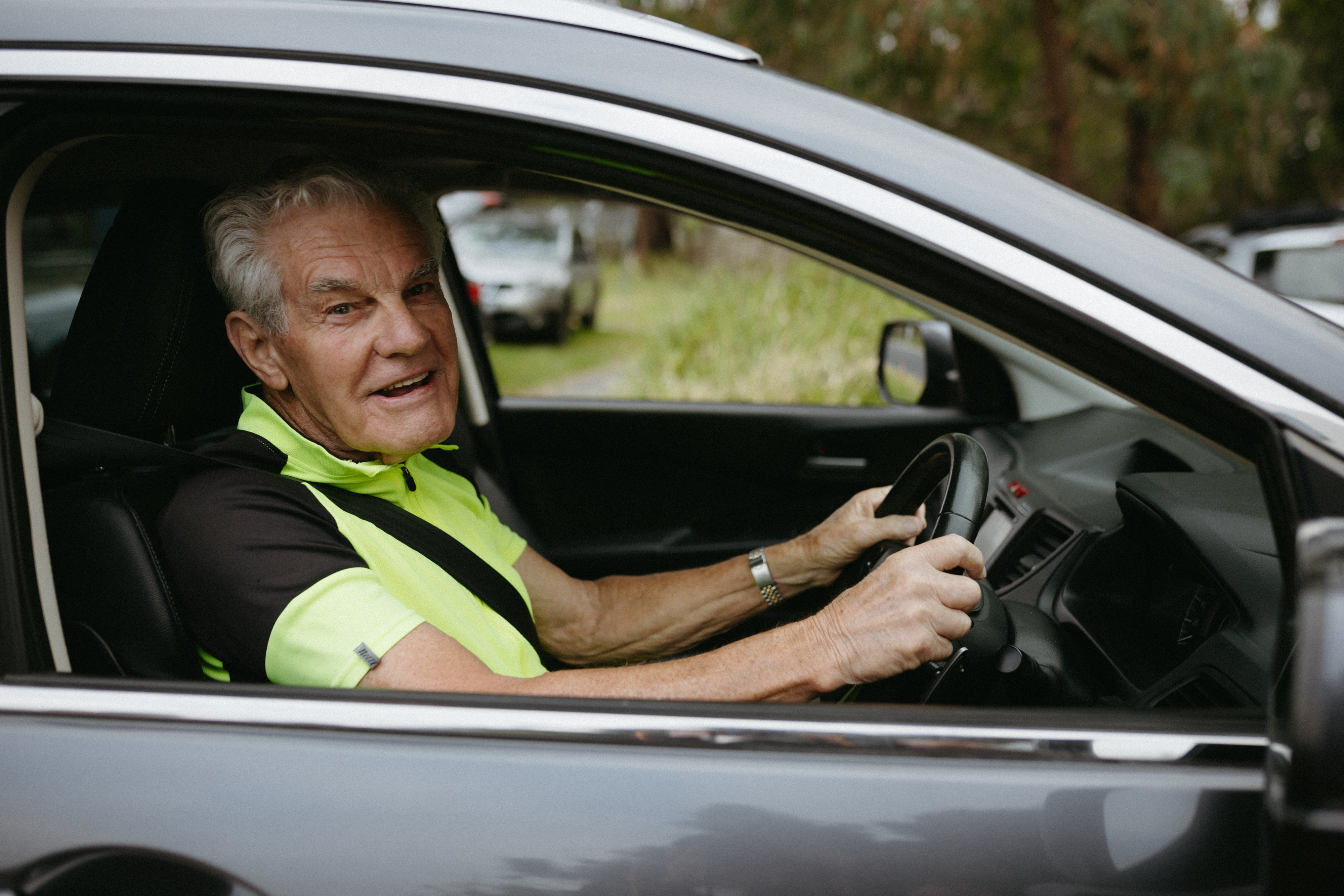 Man in his 80's seen in the front seat of a car smiling whilst wearing his bright green cycling top