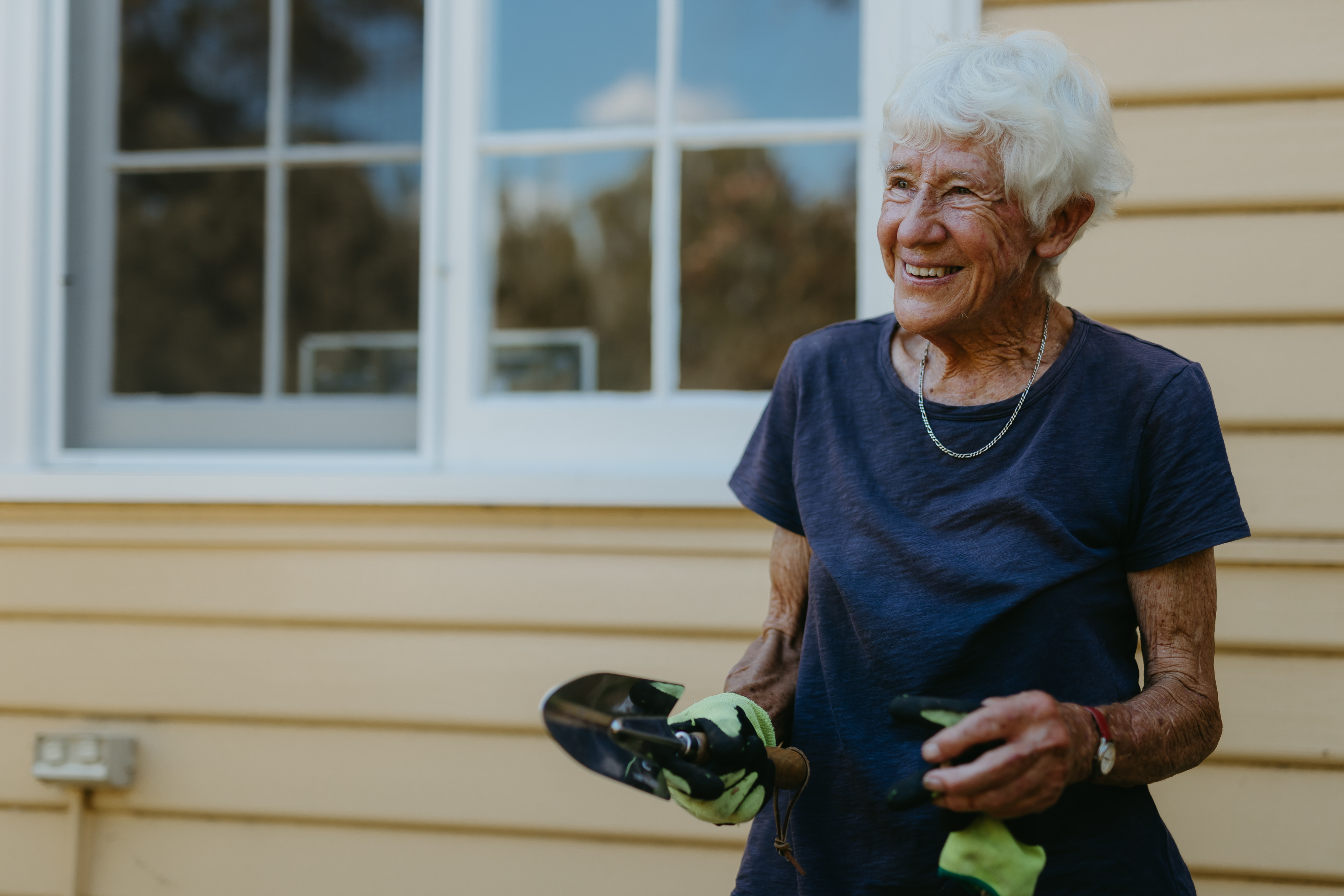 A woman in her 70s is seen smiling whilst wearing a gardening glove and holding a small shovel. 