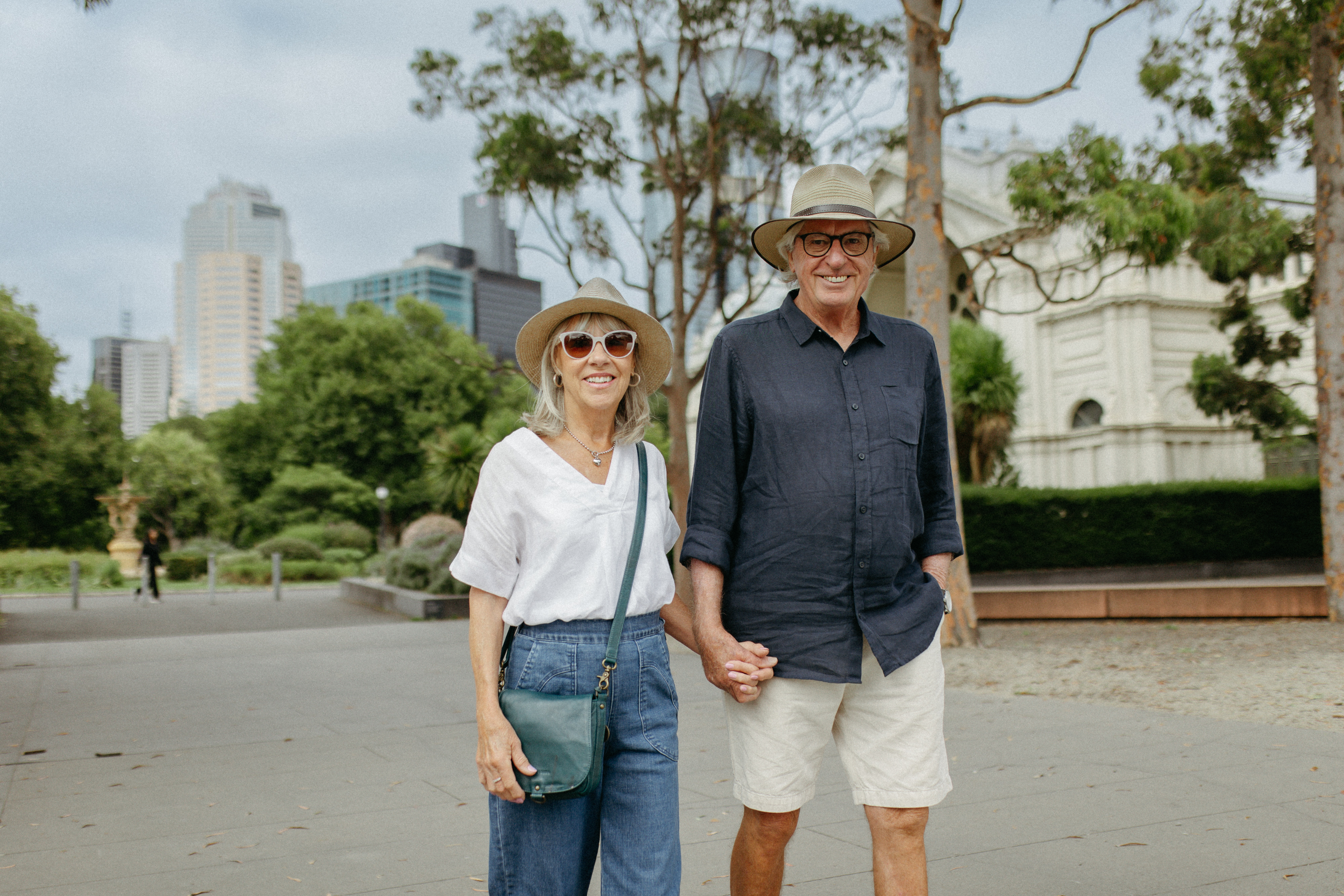 A man and lady in their 70s holding hands and walking through the green Carlton gardens with the city buildings in the background
