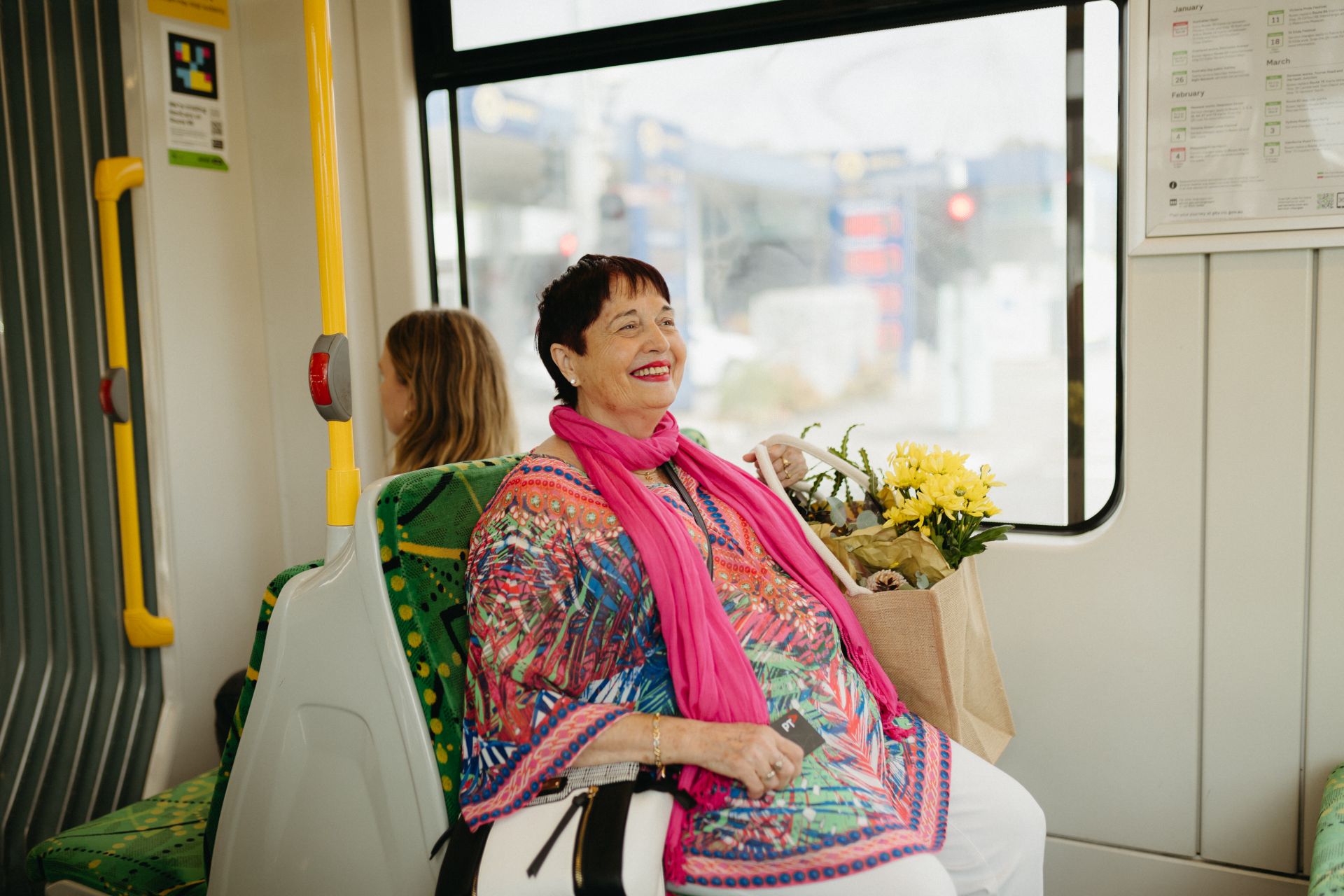 Women in bright clothing sitting on a tram seat smiling with a bag of fresh produce. 