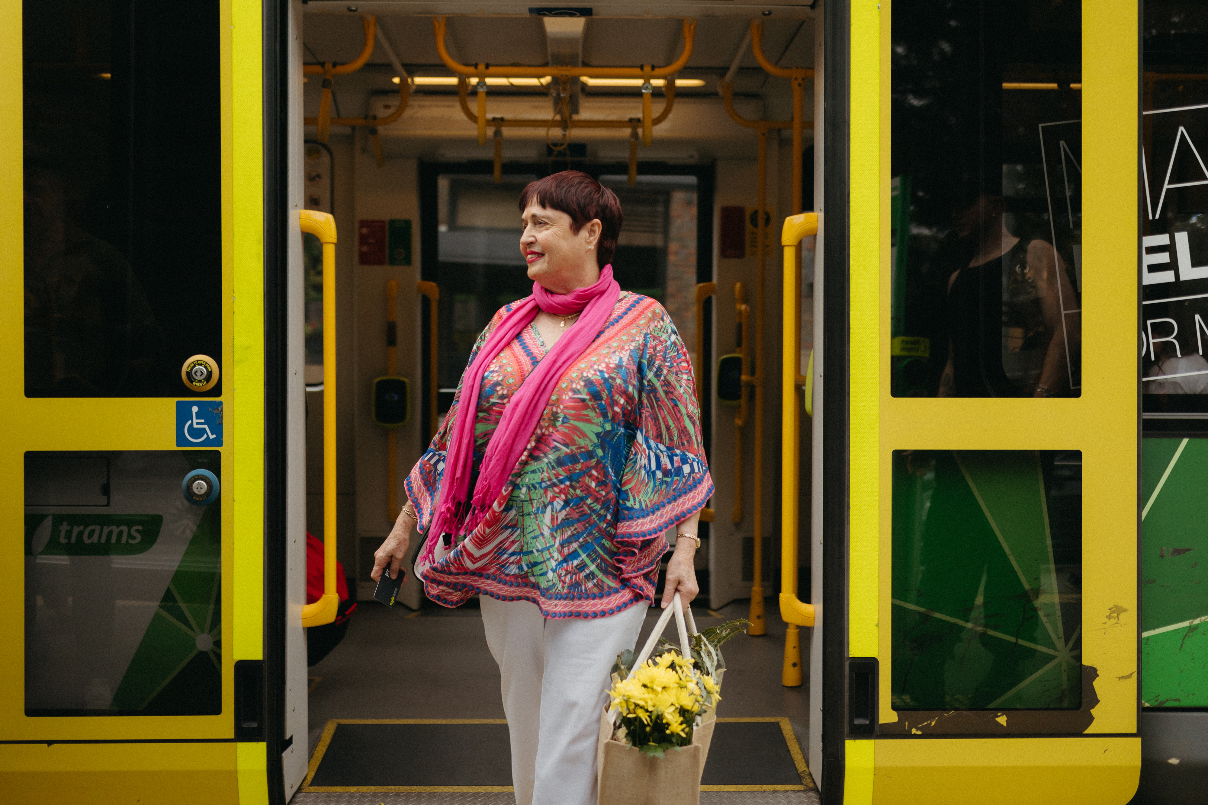 Women in bright clothing walking off the tram with a myki card in one hand and a bag of produce in the other. 