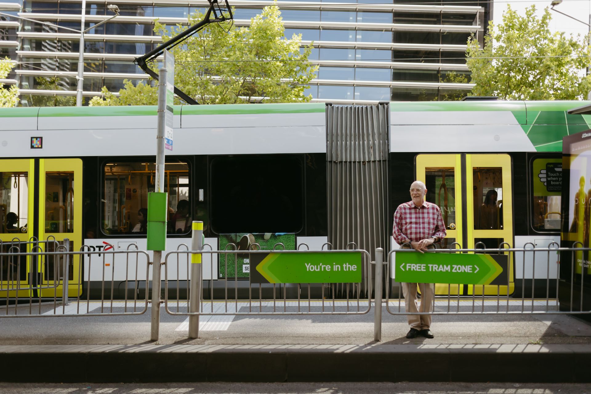 Main in red shirt standing behind the barrier at a tram strop with a tram stopped behind him