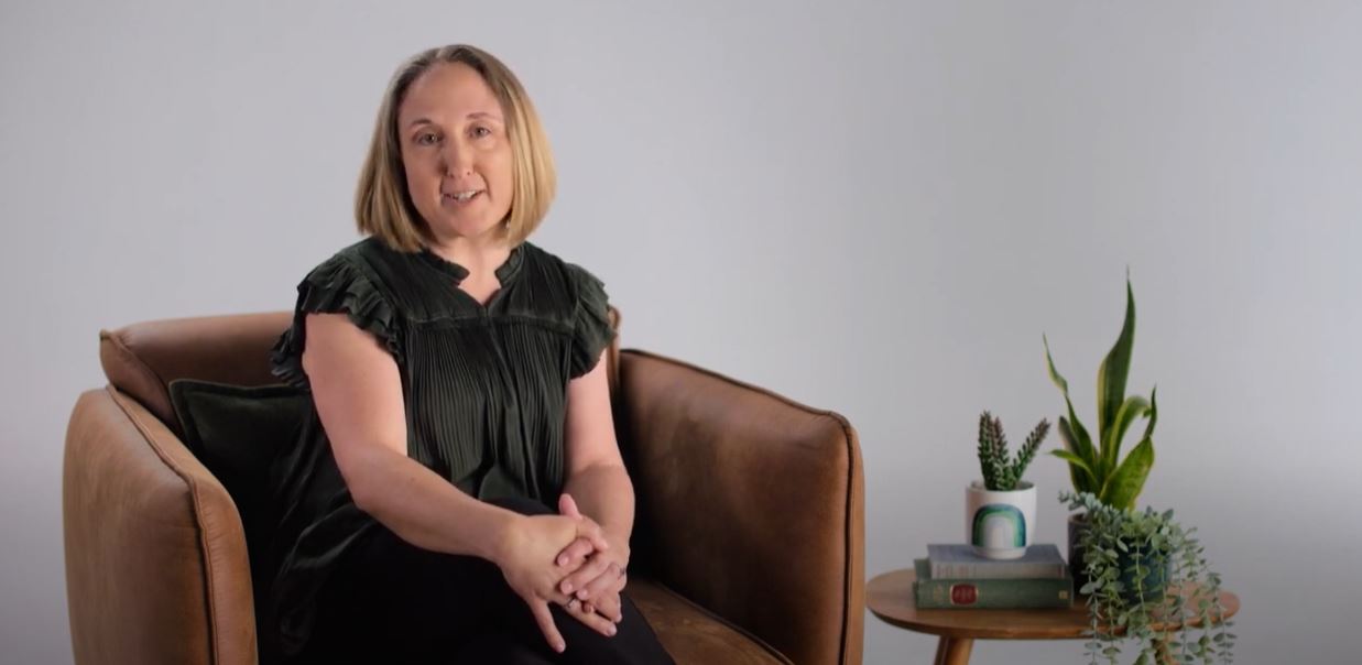 Image of a woman with a short bob wearing a dark green top sitting on a brown armchair 