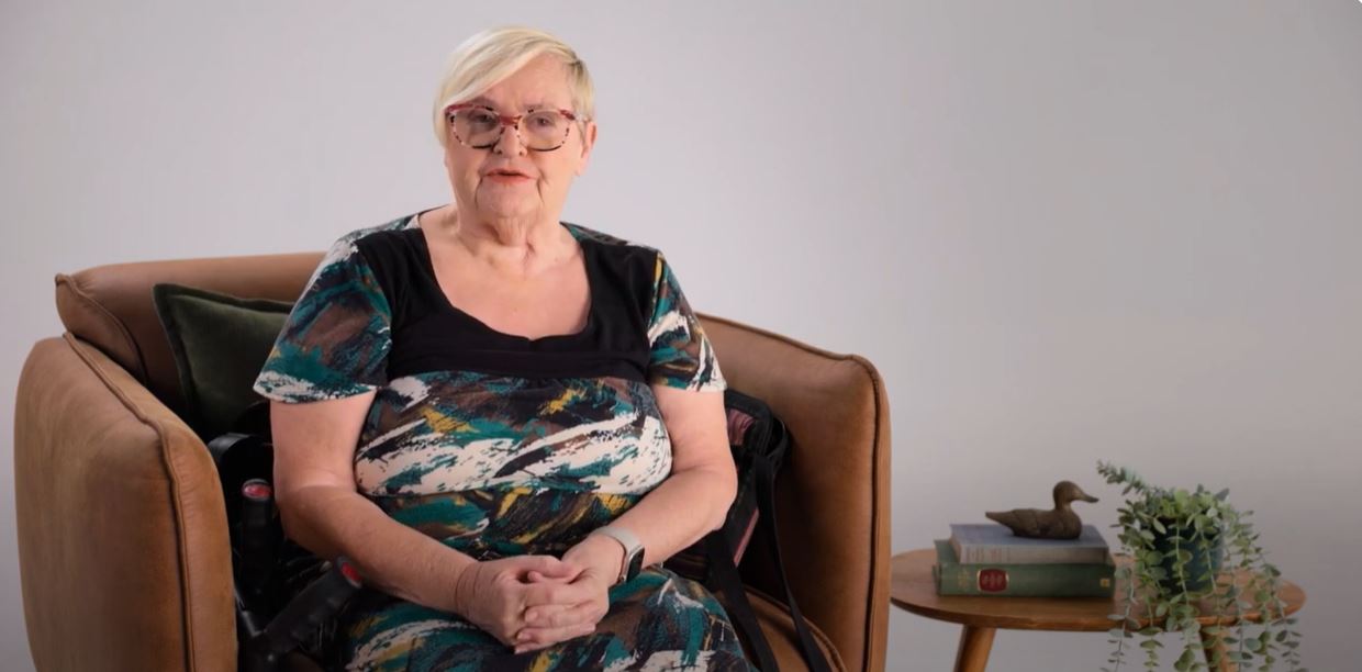 Image of a woman wearing glasses and a dress sitting on a brown armchair with her hands crossed. 